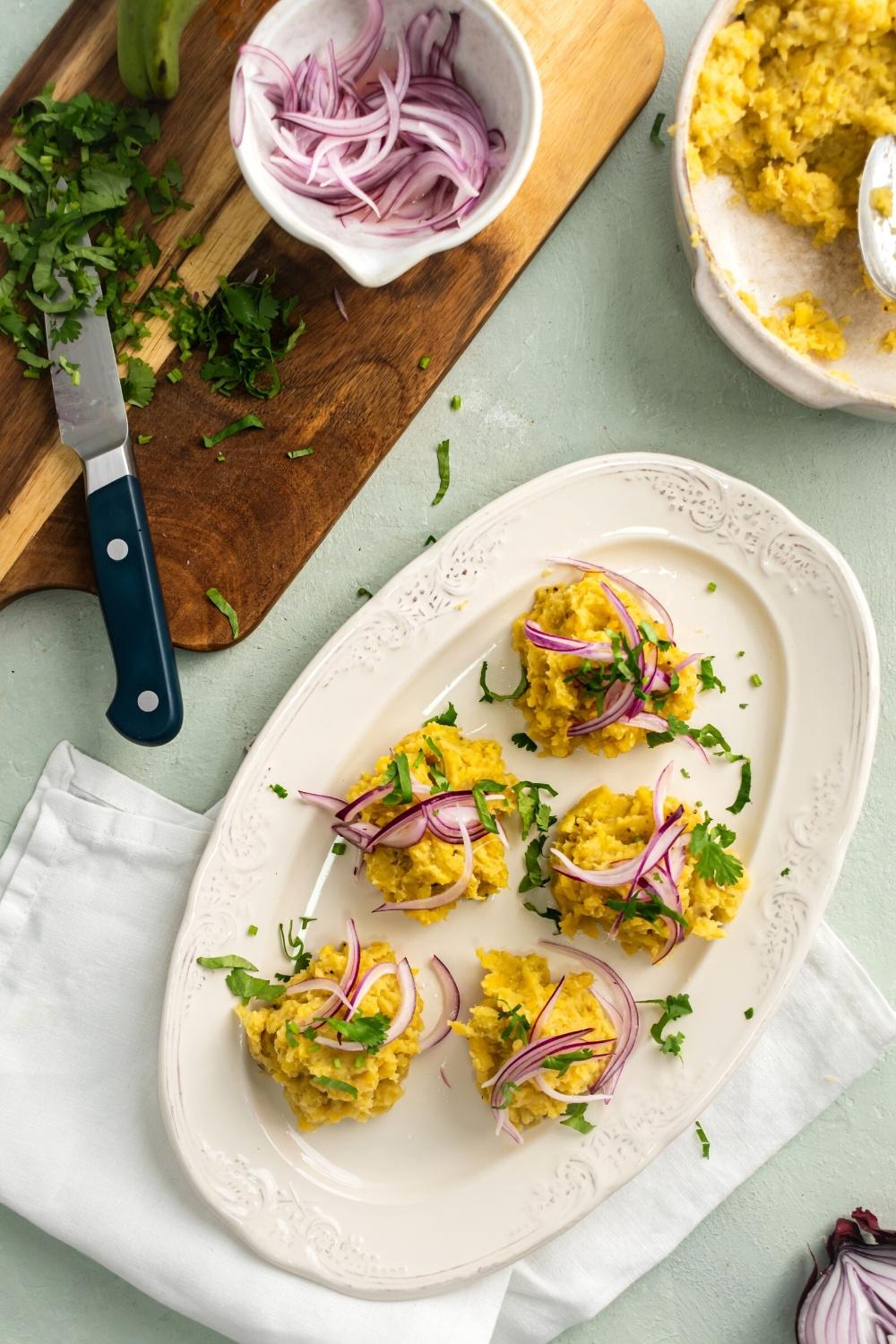 Pickled red onion on top of five scoops of mangu on a white plate that is on top of a white tablecloth on a gray counter. Behind the plate of mangu is part of a white bowl filled with the mangu and a wooden cutting board with chopped parsley and pickled red onion in a white bowl.