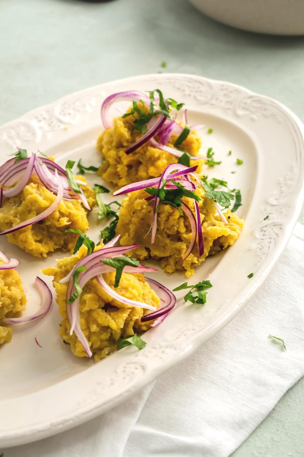 Four clumps of mashed plantains with chopped parsley and pickled red onion on top of it on a white plate.