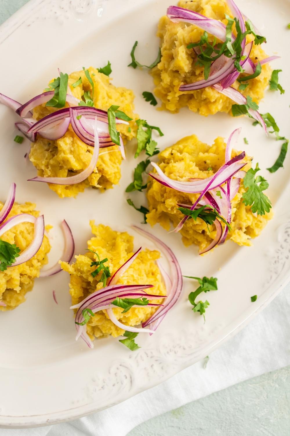 Pickled red onion and chopped parsley on top of five scoops of mangu that is on a white plate.