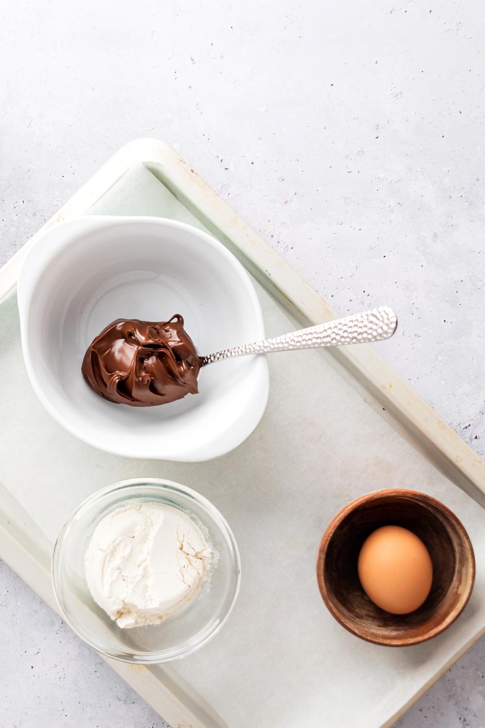 A bowl with an egg in it, a glass bowl with flour in it, a white bowl with a spoon in it that is covered with Nutella all on a piece of parchment paper on a baking sheet on the white counter.