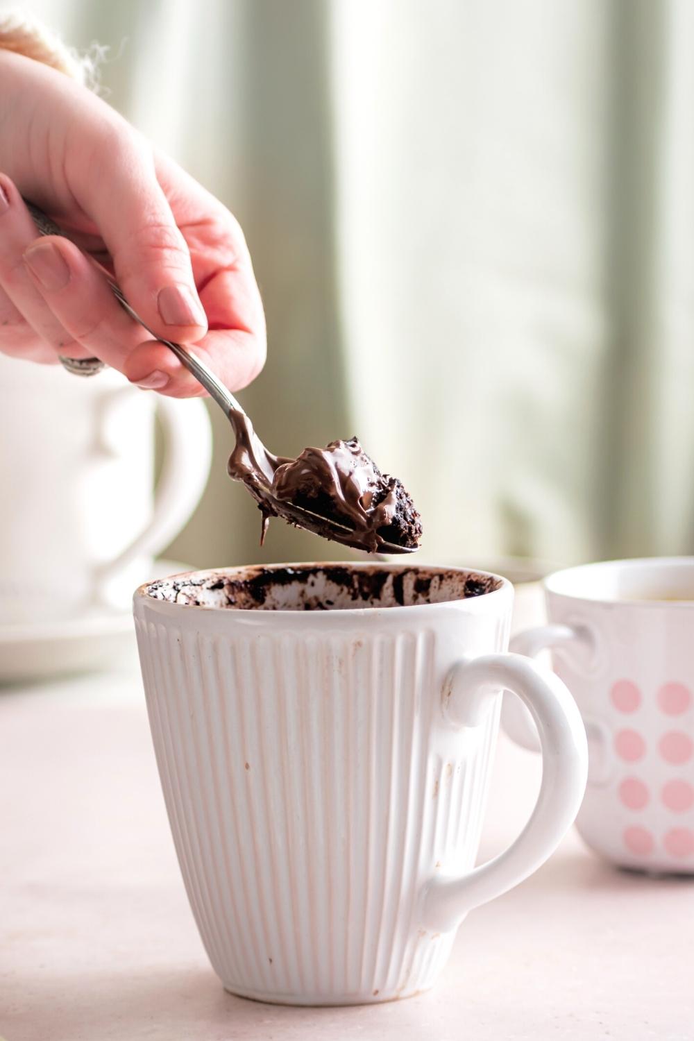 A hand holding a spoon that has a scoop of Nutella mug cake on it over a white mug that is filled with the cake.