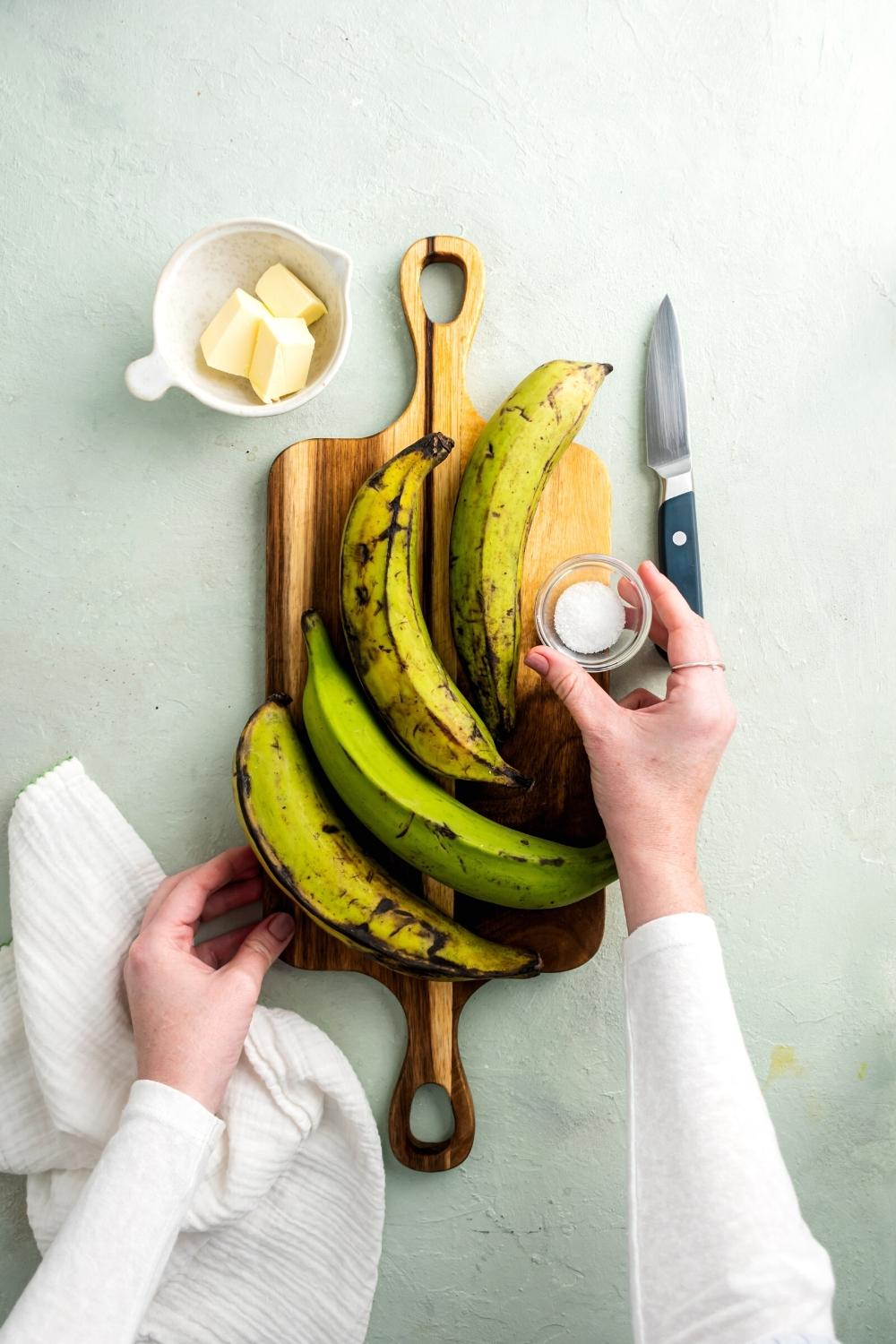 For plantains and a hand holding a glass cup of sugar on a wooden cutting board on top of a gray counter. To the right of the cutting board is a knife and to the left is a bowl of cubed butter.