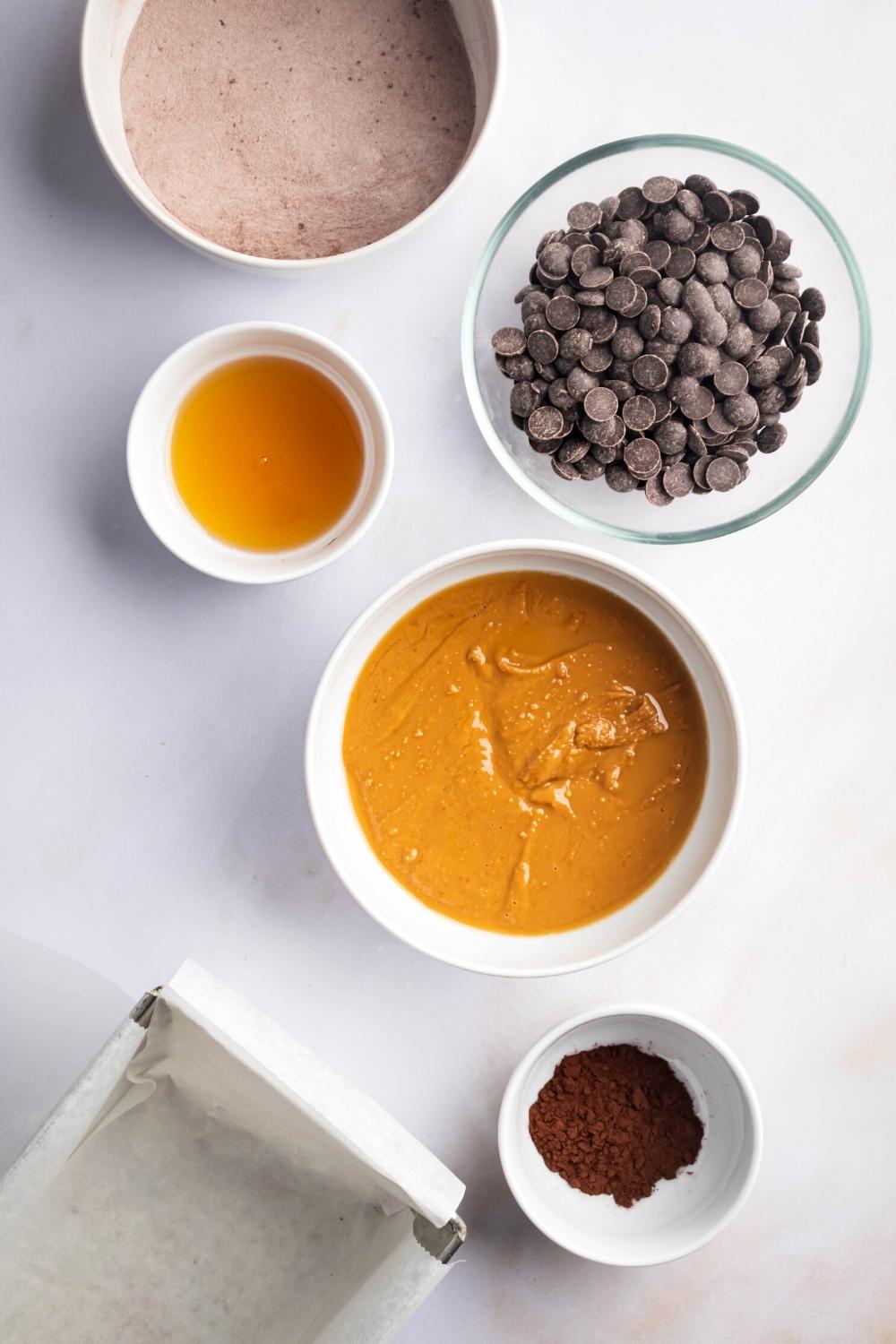 A bowl of protein powder, a bowl of chocolate chips, a small bowl of maple syrup, a bowl of peanut butter, and a small bowl of cocoa powder all on a white counter.