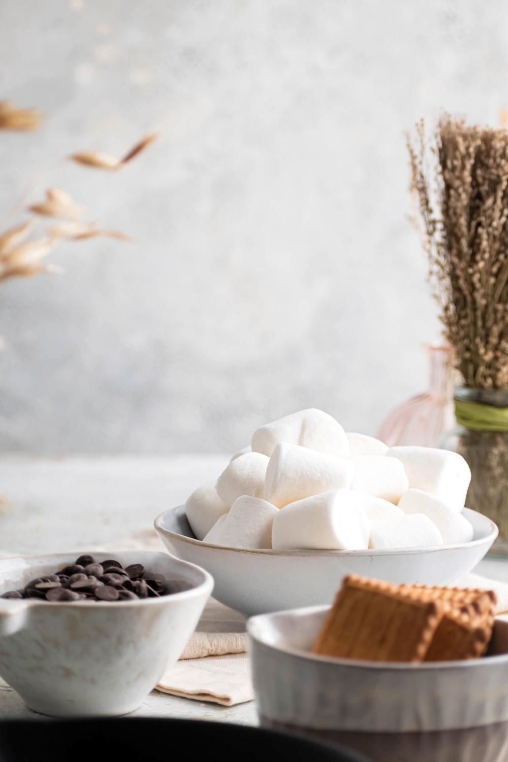 Part of a bowl of graham crackers, part of a bowl of chocolate chips, and a bowl of marshmallows on a white counter.