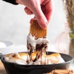 A hand holding a graham cracker that has gooey marshmallow in melted chocolate covering the bottom half of it. The marshmallow and chocolate is being pulled from s'mores dip in a cast iron skillet.