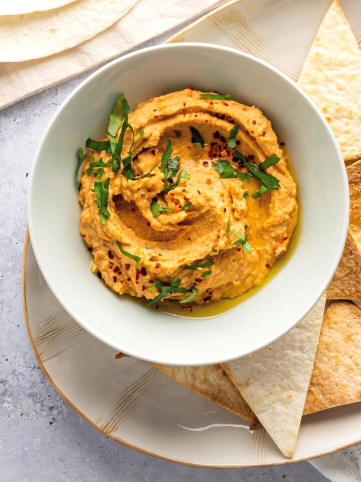 Spicy hummus in a white bowl on top of a plate that has a few tortilla chips on it's around in the bowl.