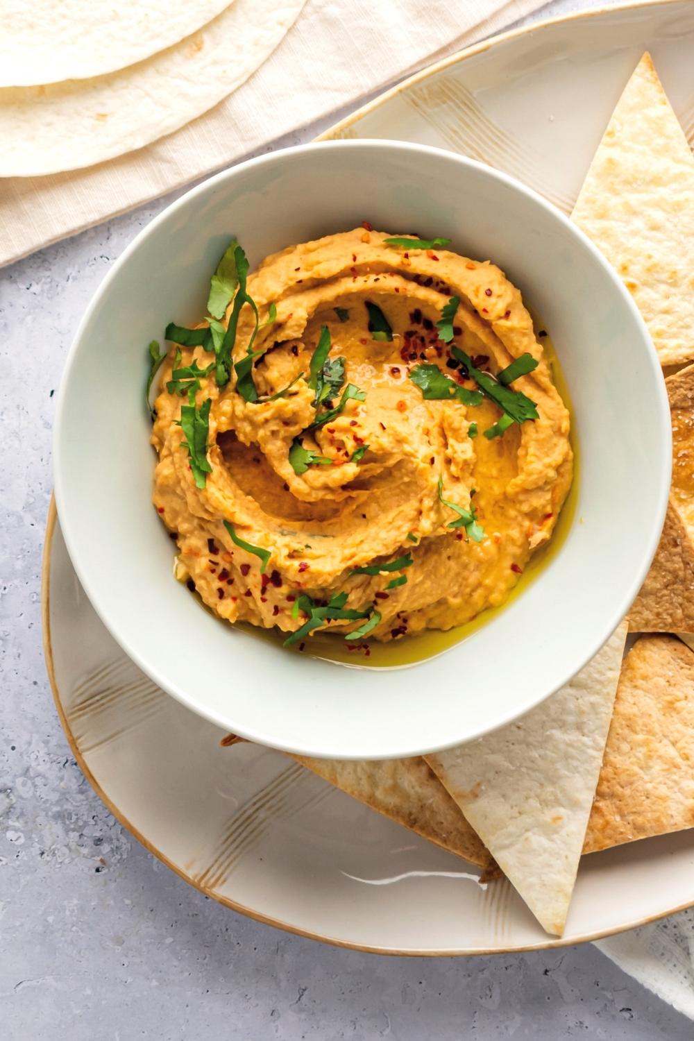 Spicy hummus in a white bowl on top of a plate that has a few tortilla chips on it's around in the bowl.