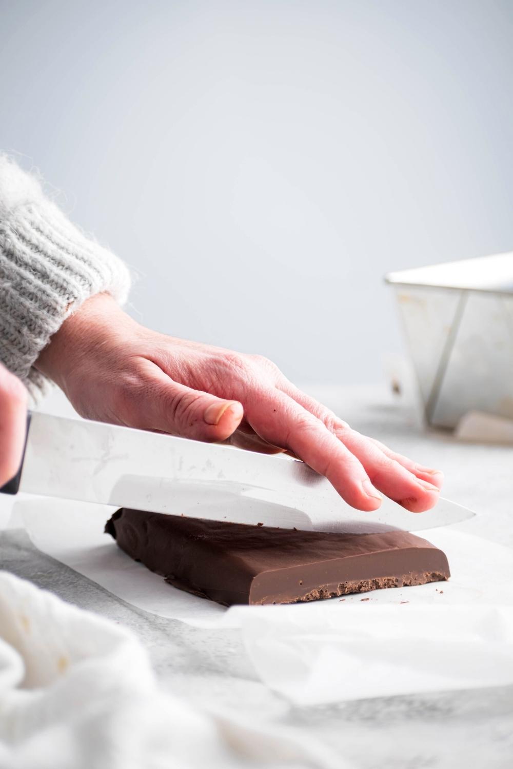 Aa hand holding a knife cutting a line of fudge out of a block of fudge on a piece of parchment paper.