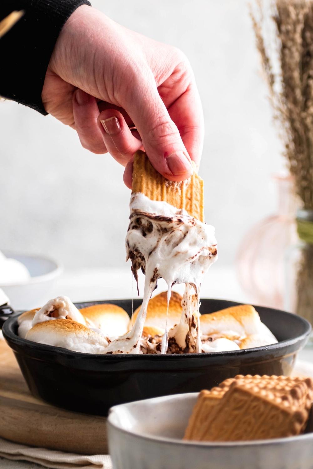 A hand holding a graham cracker that has gooey marshmallows and chocolate on it that is dripping down into a cast iron skillet filled with s'mores dip.