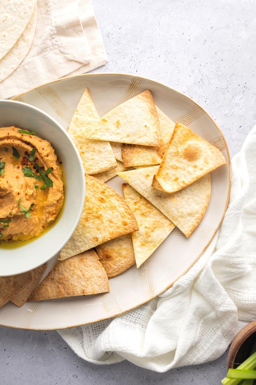 Air fryer tortilla chips on part of a plate. Next to the chips is part of a white bowl filled with hummus.