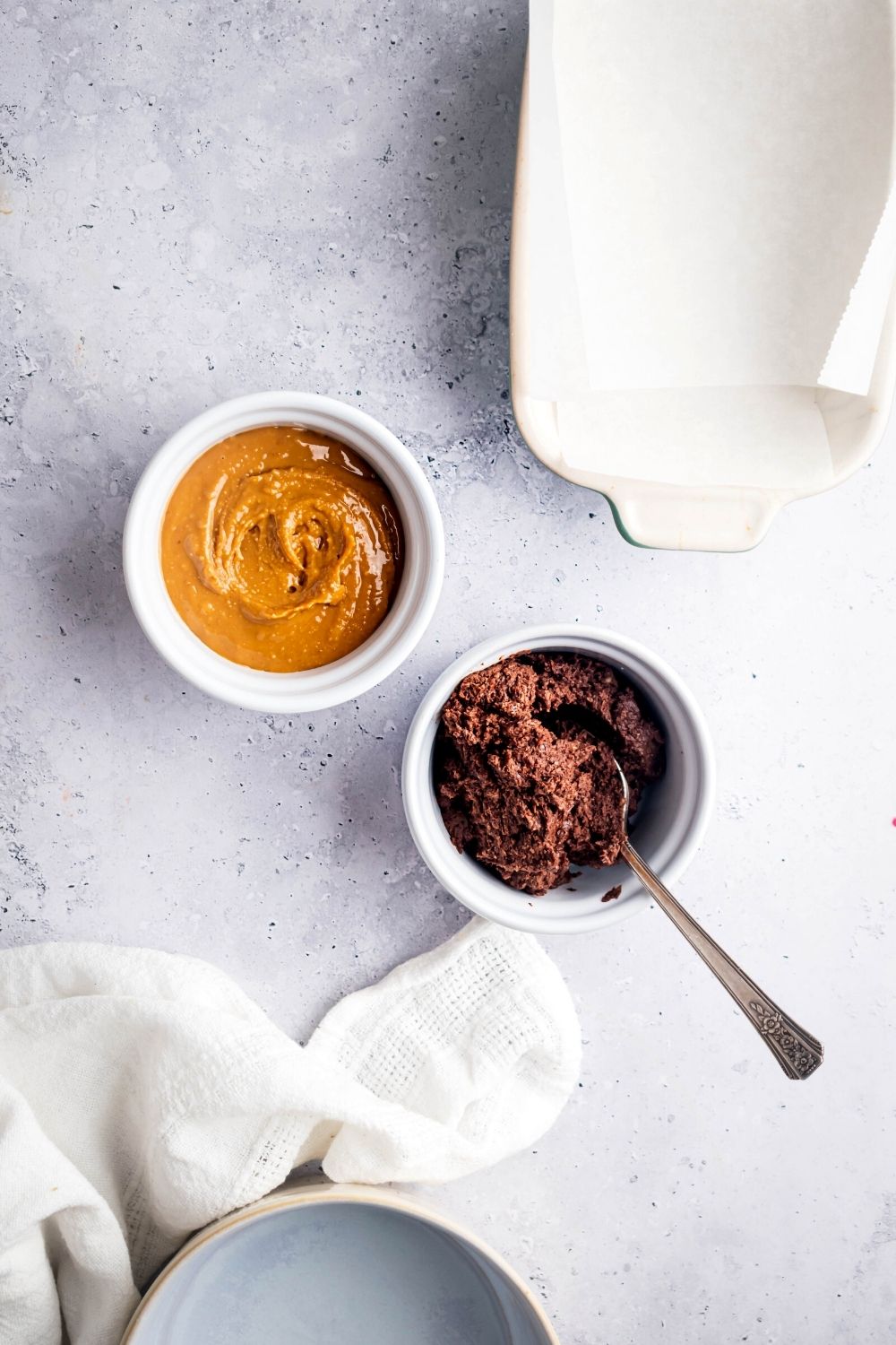 Chocolate frosting in a small white bowl, and peanut butter in a small white bowl on a white counter.