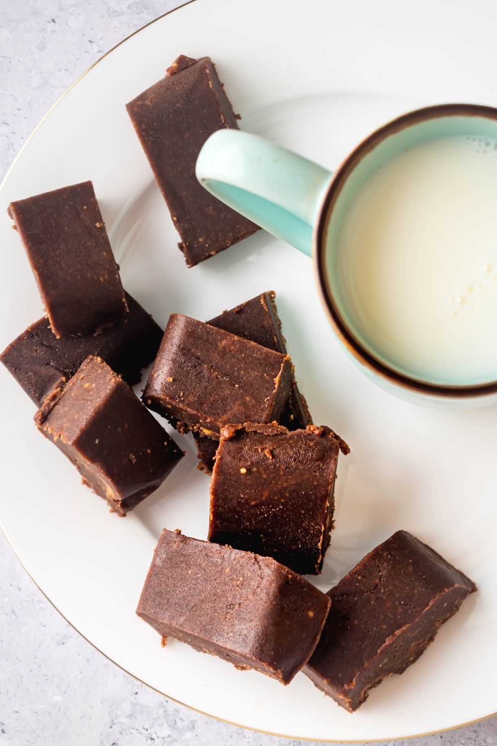 A bunch of squares of chocolate peanut butter fudge on a white plate. To the right of the pieces of fudge is part of a mug filled with milk.