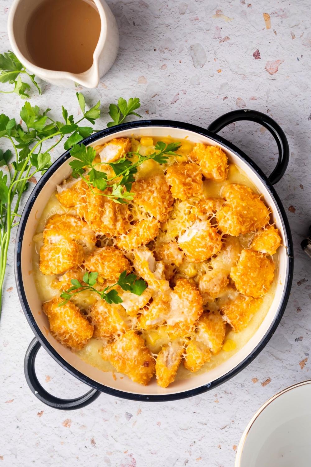 Melted shredded cheese on top of popcorn chicken and corn that is on top of mashed potatoes in a circular baking dish. Behind it is a picture of gravy and they are both on a white counter.