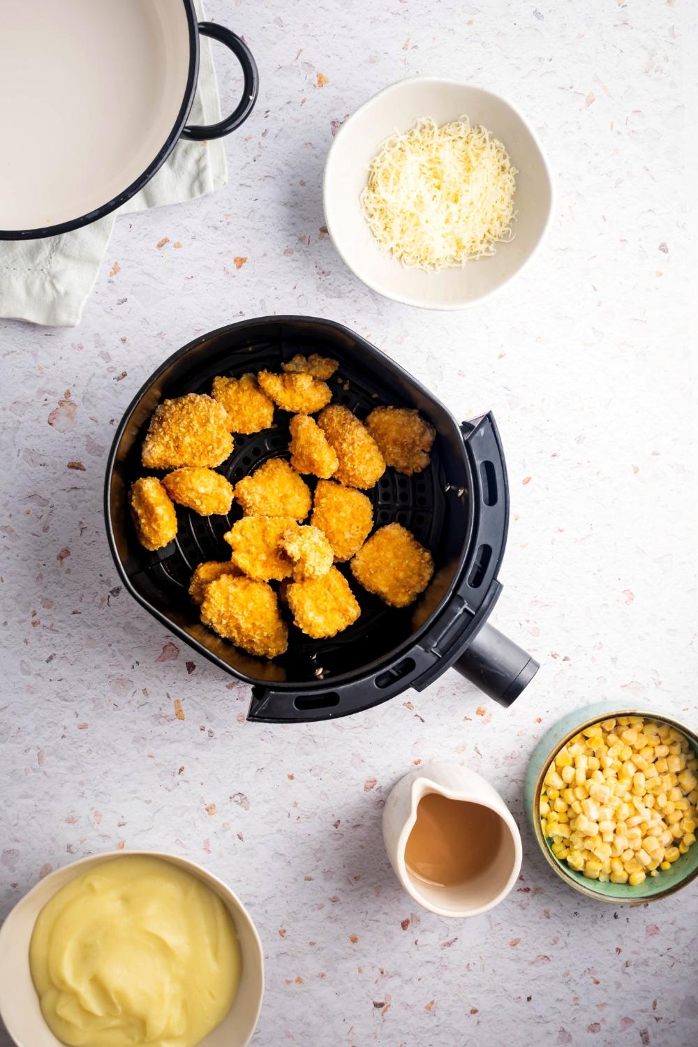 An air fryer filled with popcorn chicken surrounded by a bowl of shredded cheese, a bowl of corn, a picture of gravy, and a bowl of mashed potatoes all on a white counter.