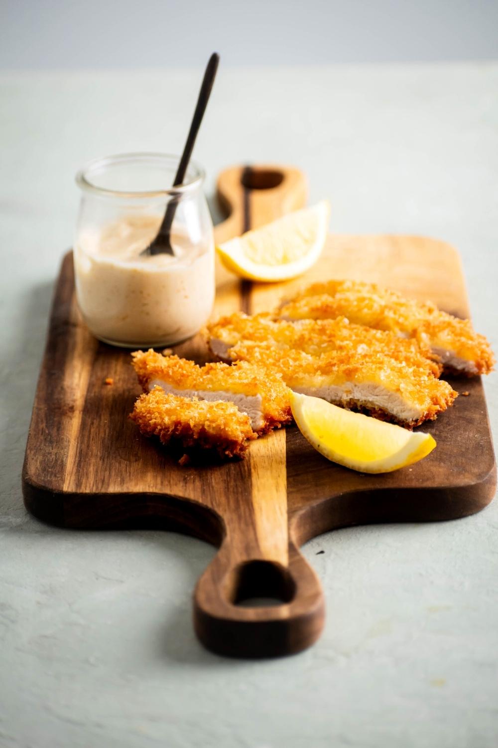 A chicken cutlet that is sliced into strips on a wooden cutting board. There are two lemon wedges in a jar of chipotle mayo on the cutting board.