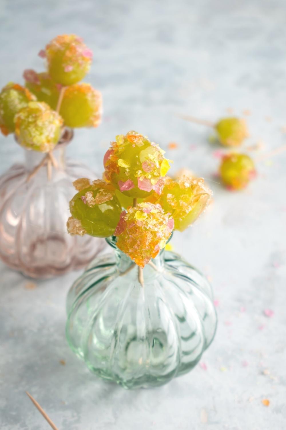 A glass vase that is filled with a bunch of candy grapes with jolly ranchers on them with toothpicks in them. Behind that is another base with the candy grapes in it.