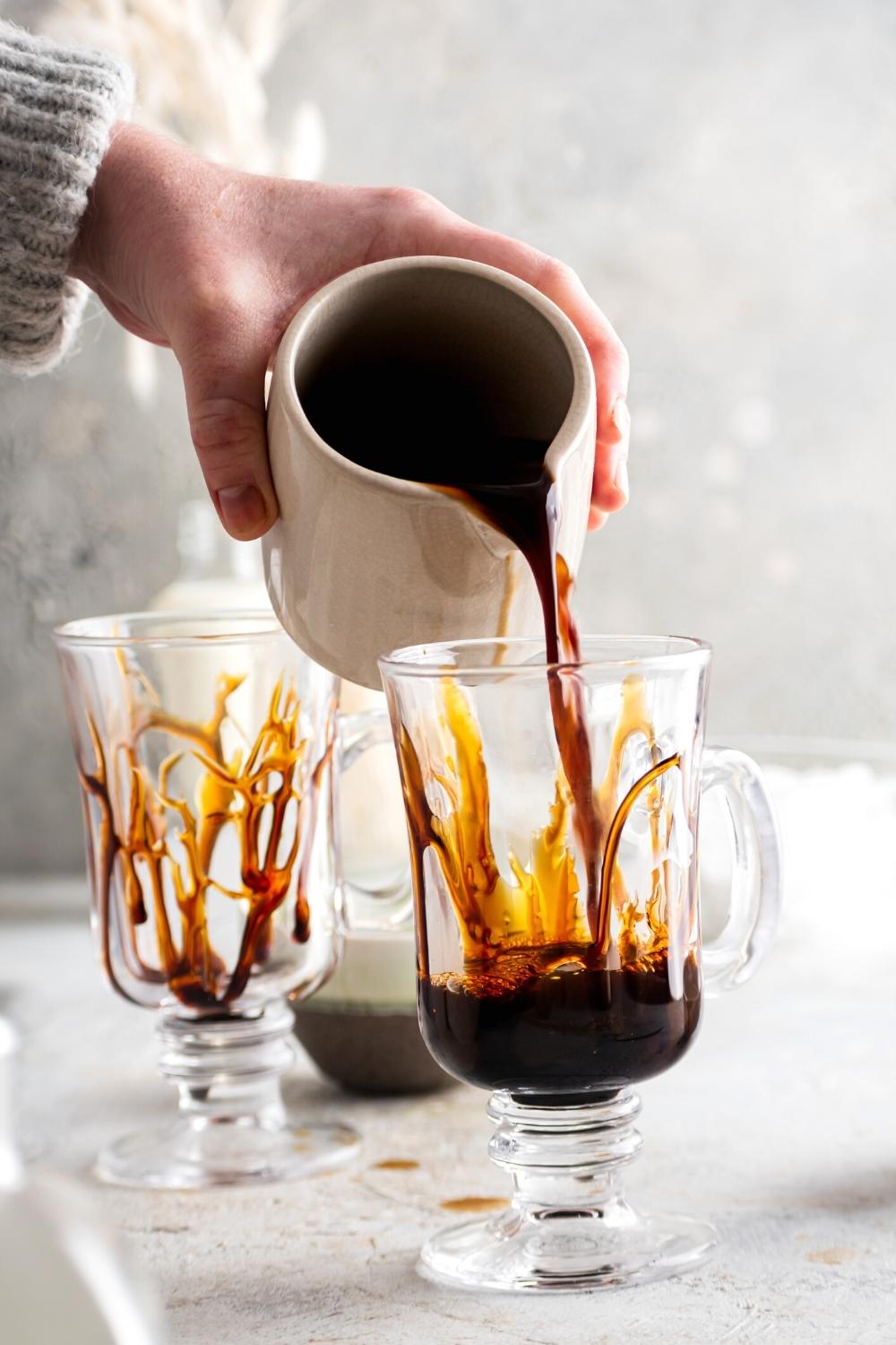 A hand holding a pitcher of caramel cold brew pouring it into a glass cup that has caramel sauce on the edges.