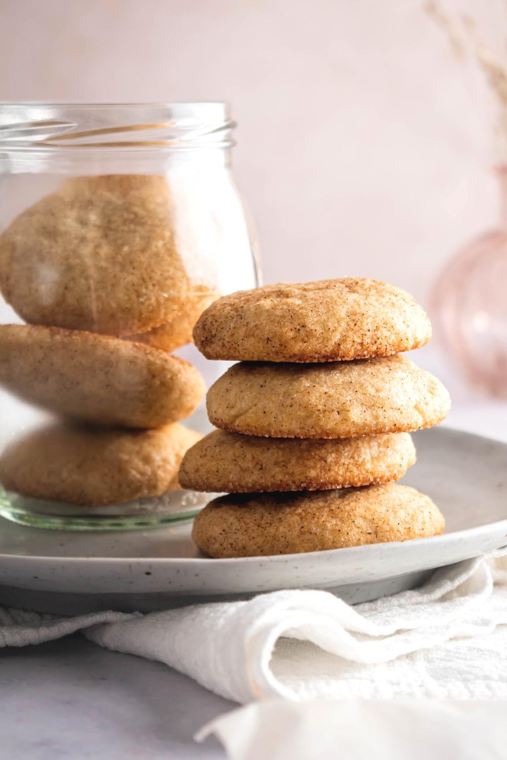 A stack of four snickerdoodle cookies on a white plate. Next to the stack as a jar filled with snickerdoodle cookies.