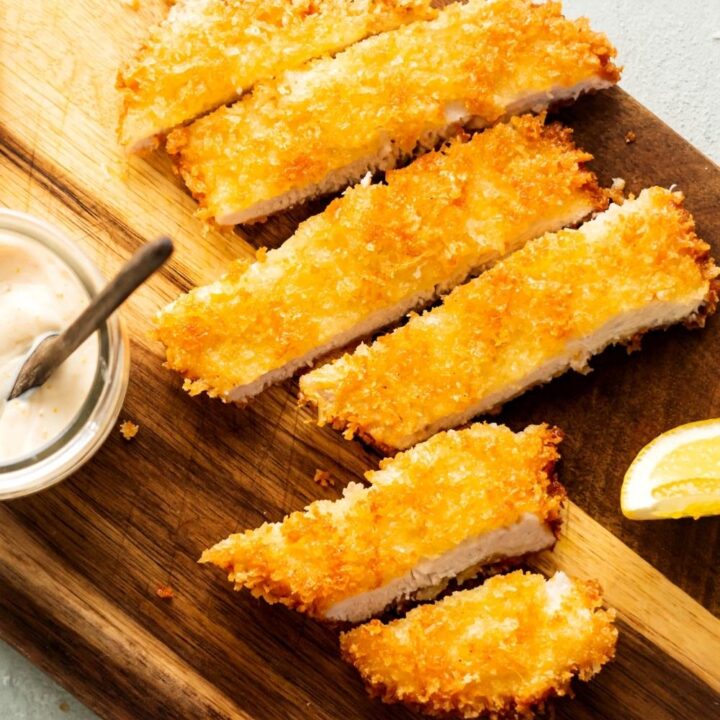 Six slices of breaded chicken cutlet on a wooden cutting board.