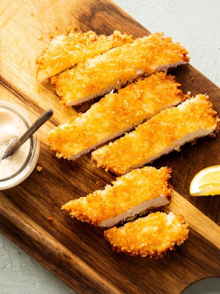 Six slices of breaded chicken cutlet on a wooden cutting board.