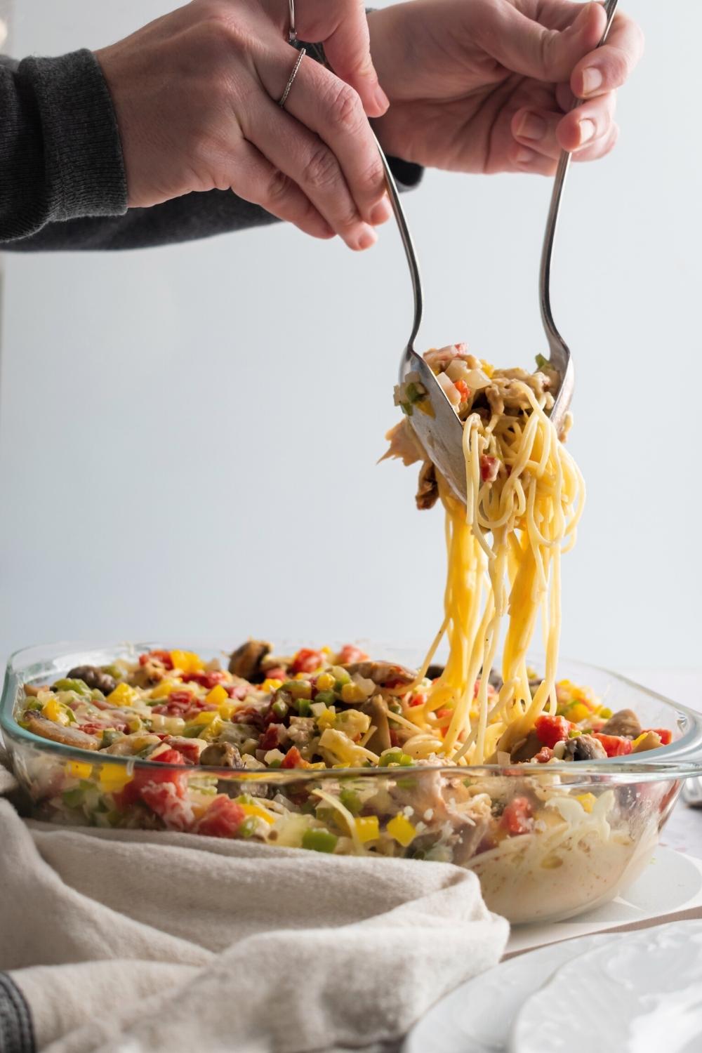 Two hands holding utensils with spaghetti between them, hovering over a glass casserole dish with the Texas chicken spaghetti in it.
