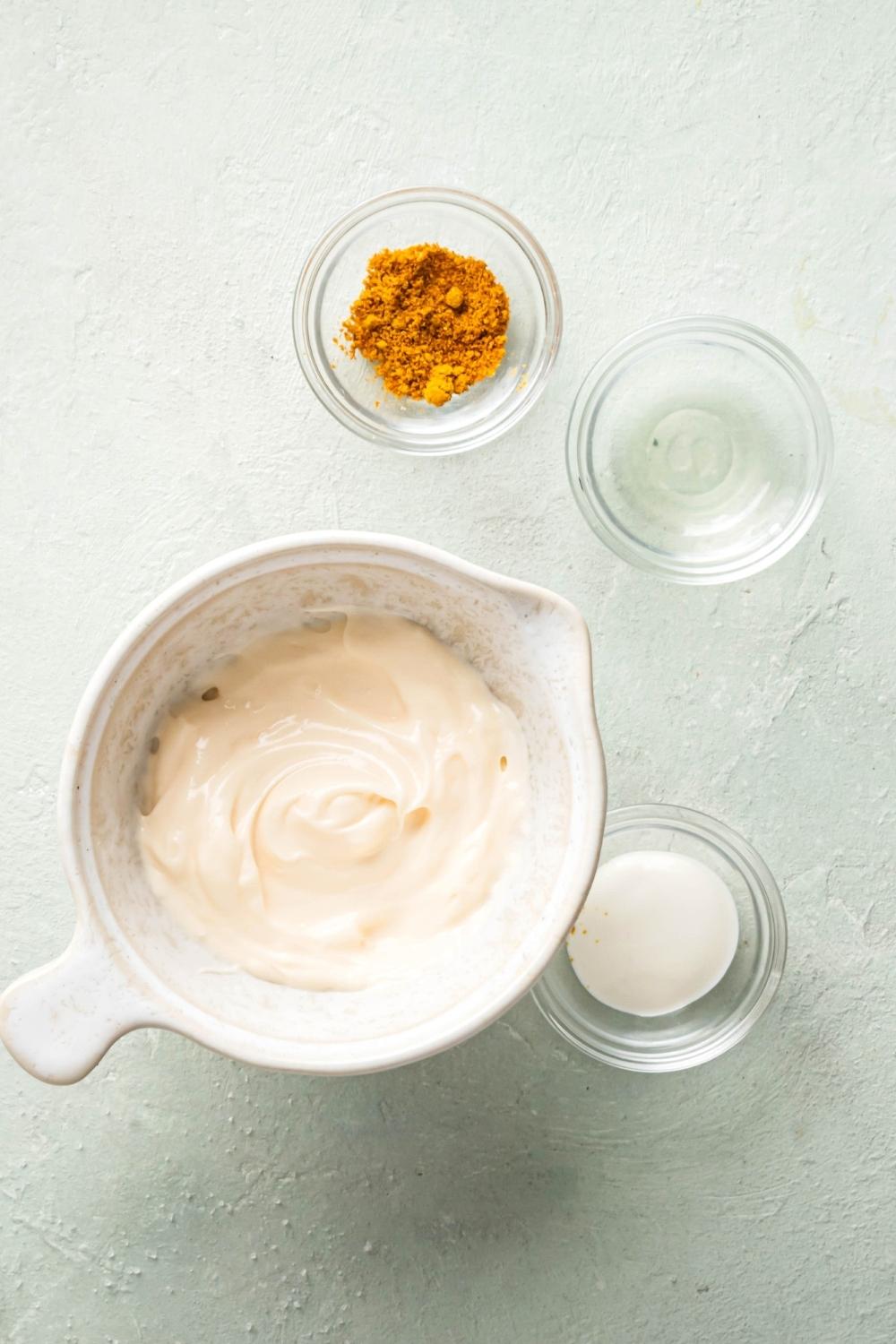 A bowl of chipotle mayo, a bowl of sugar, and empty glass bowl, and a bowl of chipotle powder all on a white counter.