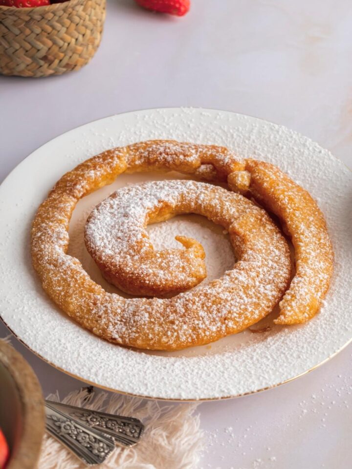 A fried funnel cake with powdered sugar on top on a white plate on a white counter.