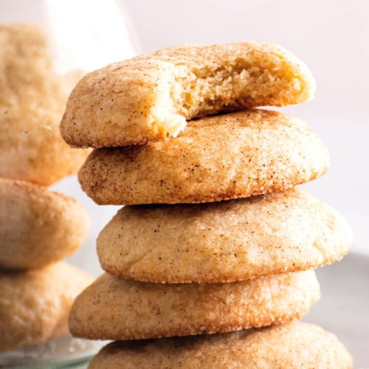 Five snickerdoodles stacked on top of one another on a white plate. The cookie on the top has a bite out of the front of it.