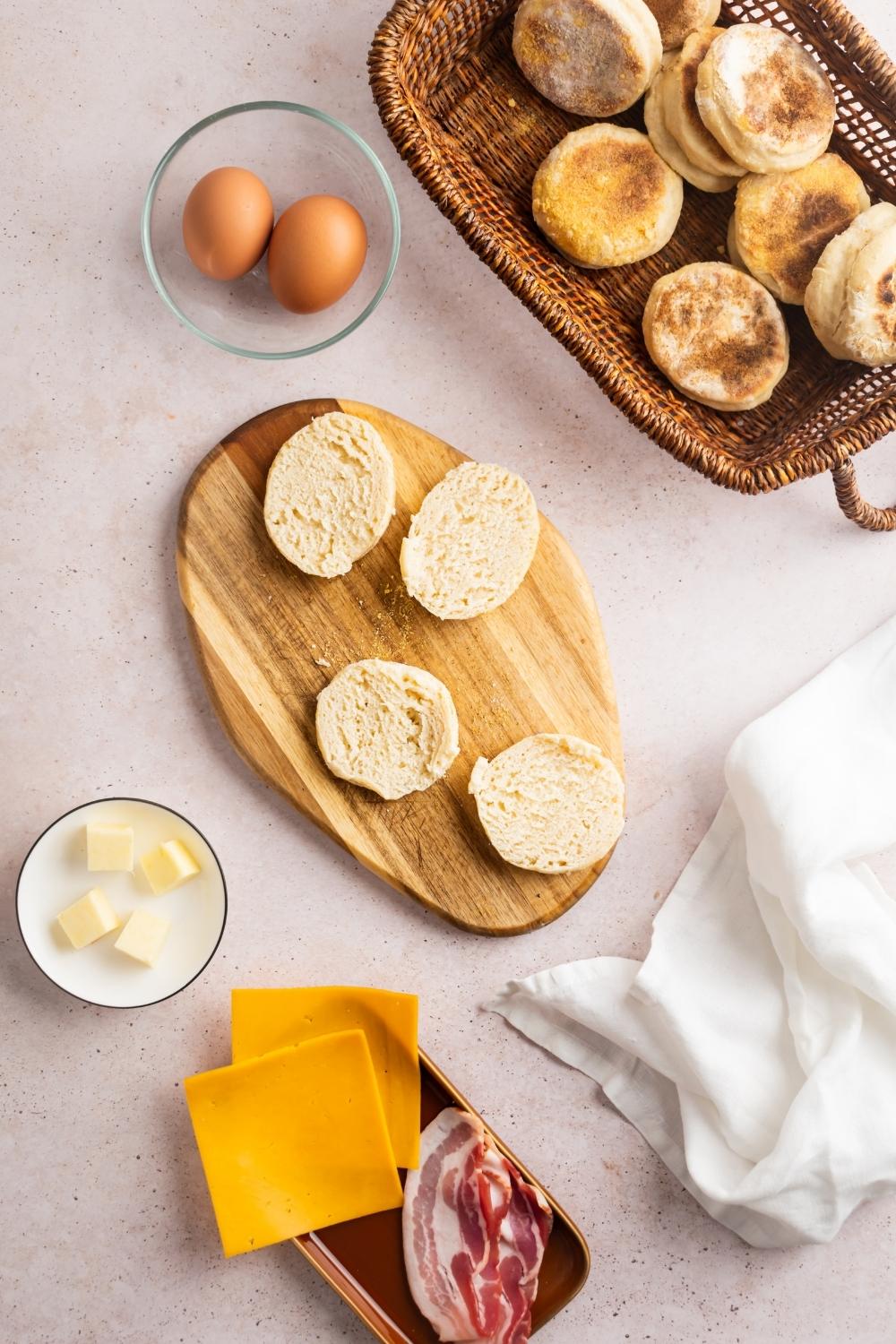 Two opened up english muffins on a wooden cutting board, butter cubes in a bowl, two eggs in a bowl, a basket of english muffins, and two slices of cheese in a bowl all on a white counter.