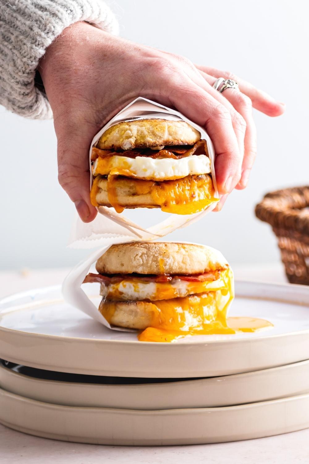 A hand holding an egg mcmuffin above another egg mcmuffin wrapped in parchment paper.