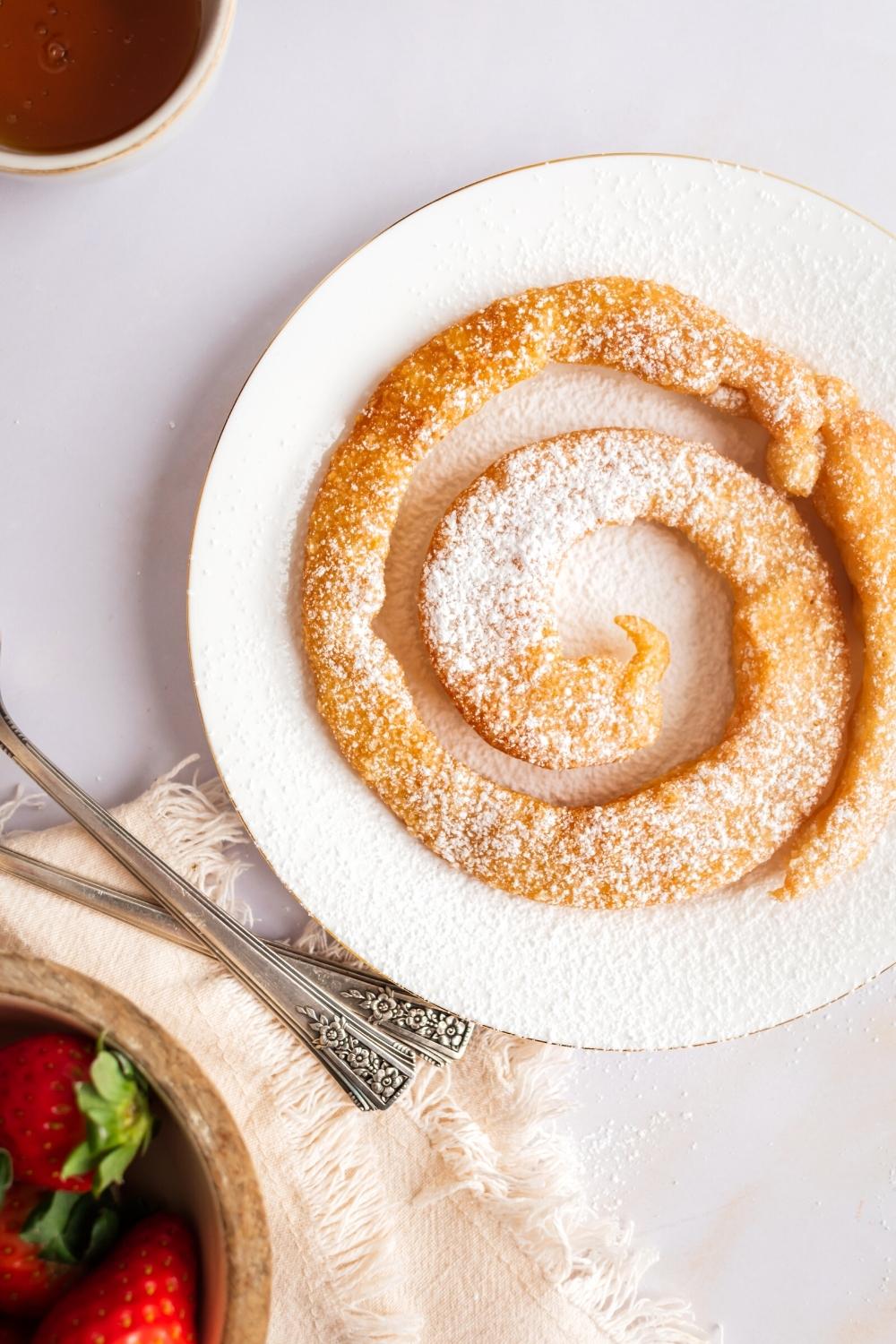 Powdered sugar on top of a circular funnel cake on a white plate on a white counter.