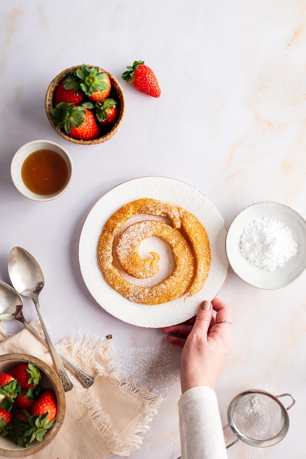 A hand holding a plate on a white counter that has a circular funnel cake on it. Surrounding the plate is a bowl of powdered sugar, a bowl of strawberries, and a bowl of syrup.