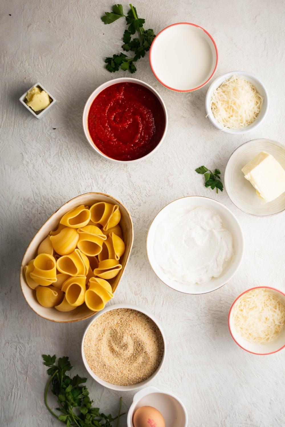 Bowl of breadcrumbs, a bowl of ricotta cheese, of all with butter, ball shredded cheese, bowl marinara sauce, and a bowl of giant shells on a white counter.