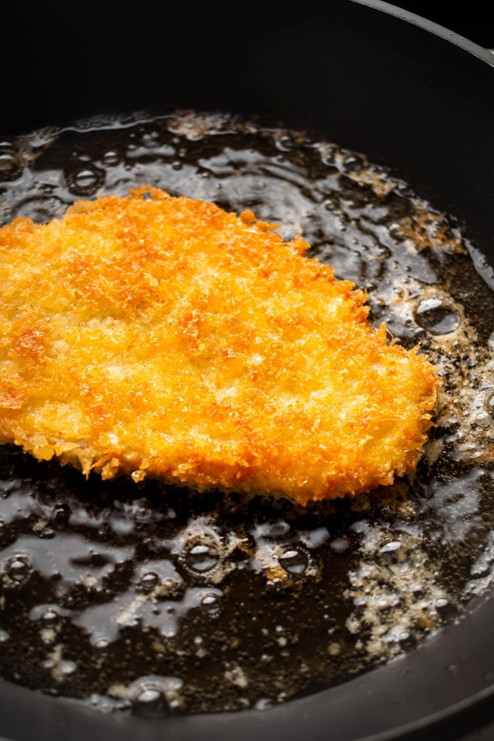 A breaded chicken cutlet that is cooked to Golden perfection in oil in a pan.