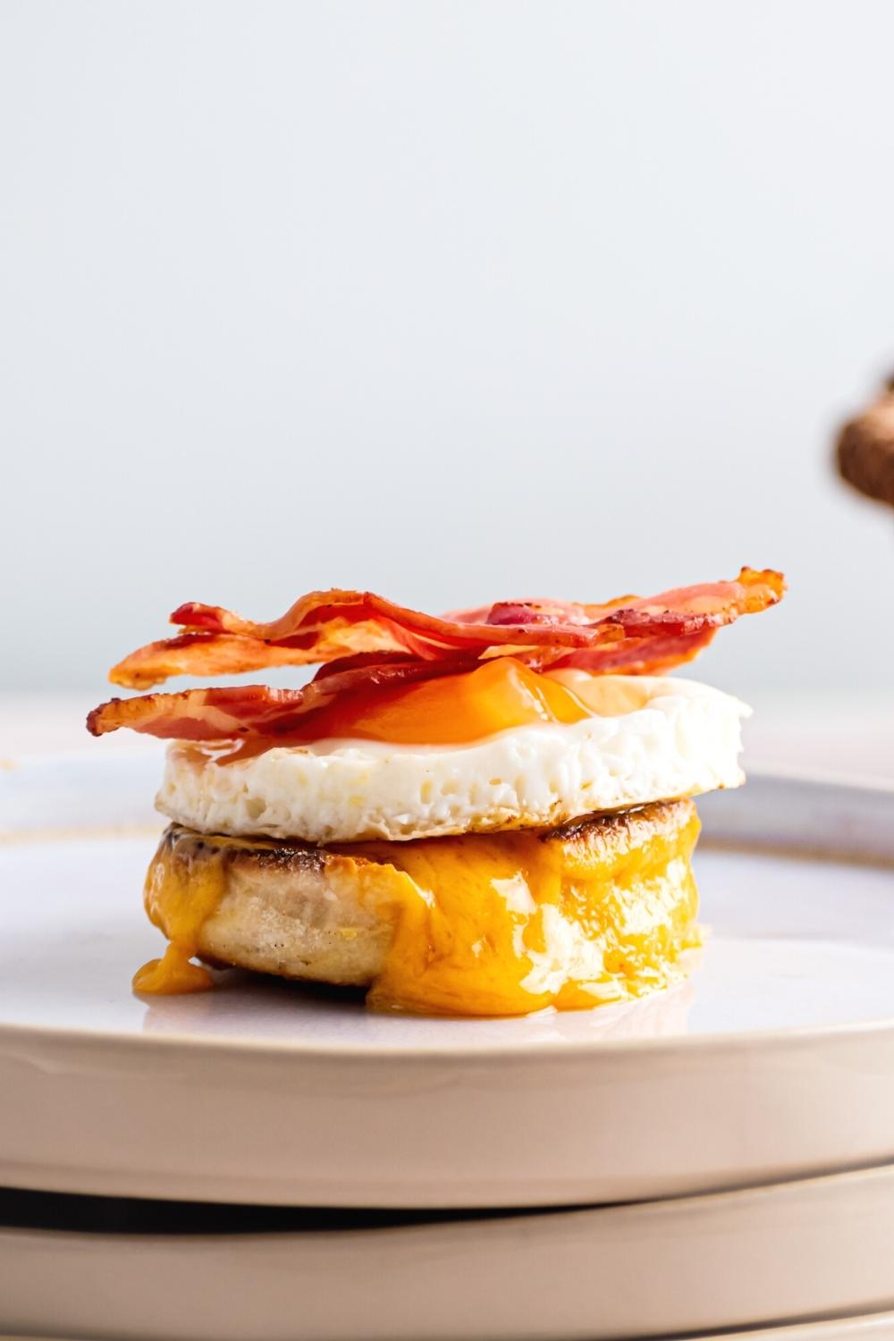 Two slices of bacon on an egg on top of cheese on an English muffin on a plate.