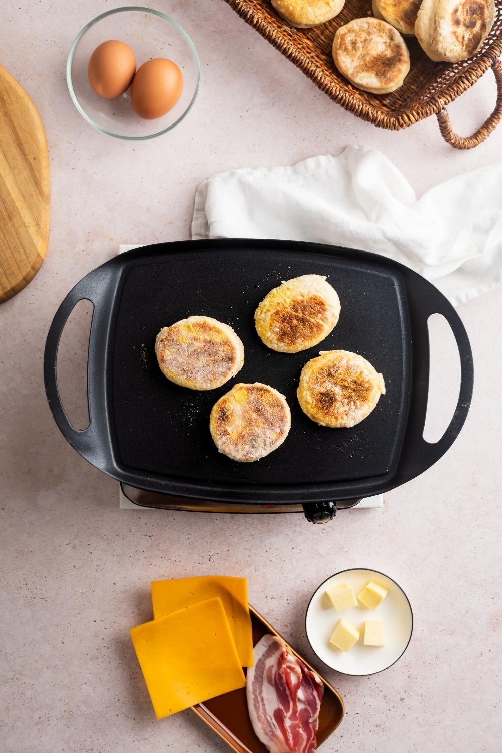 Two english muffins face down on a griddle. Surrounding it is two eggs in a bowl, part of a basket with english muffins, and a bowl with two slices of American cheese all on a white counter.