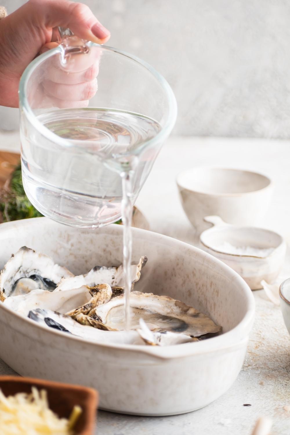 A hand pouring boiling water into an oyster shell. The oyster is in a baking dish with six other oysters.