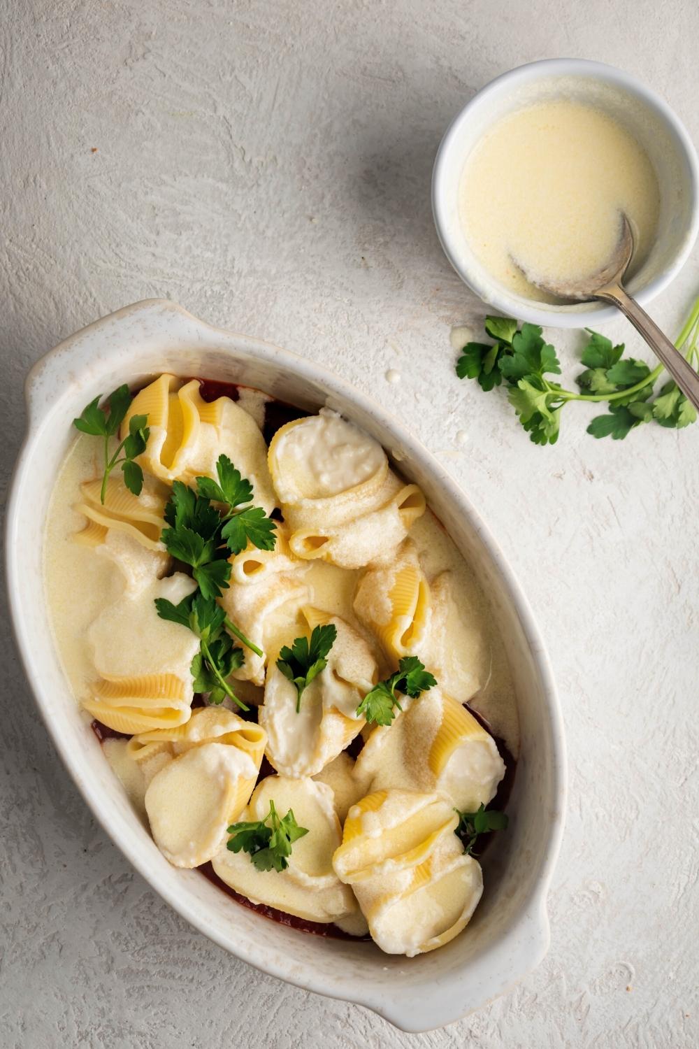 Giant cheese stuffed shells with Alfredo sauce on them in a white casserole dish. Behind it is a white bowl with Alfredo sauce in it.