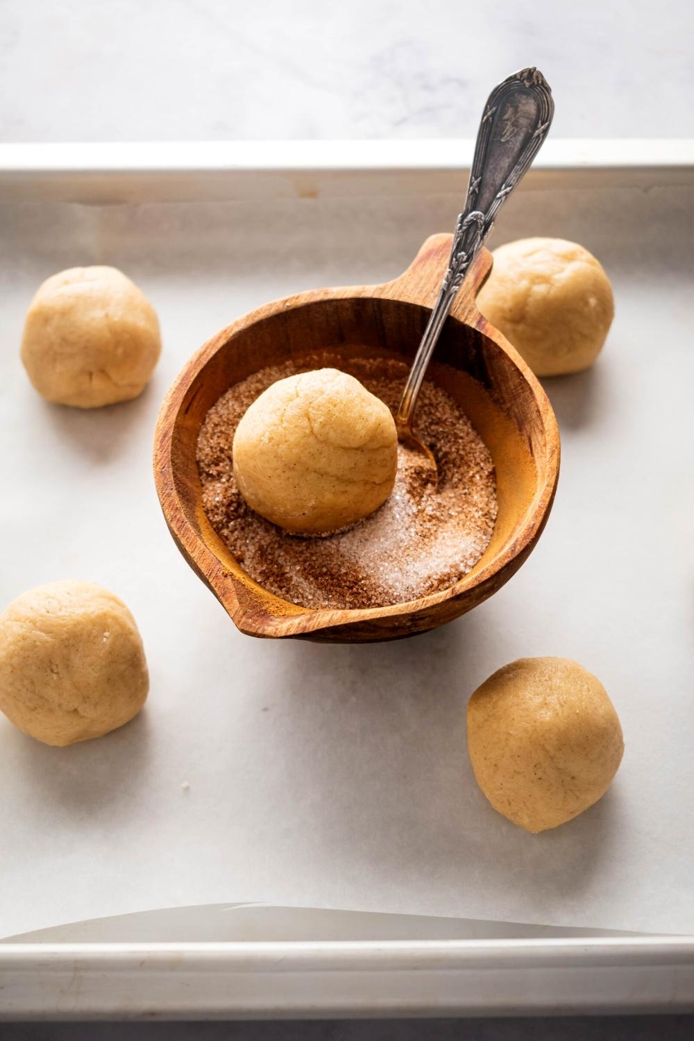 A snickerdoodle cookie dough ball in a ball that is filled with cinnamon and sugar. The ball is on top of a piece of parchment paper that has four snickerdoodles on it surrounding the bowl.