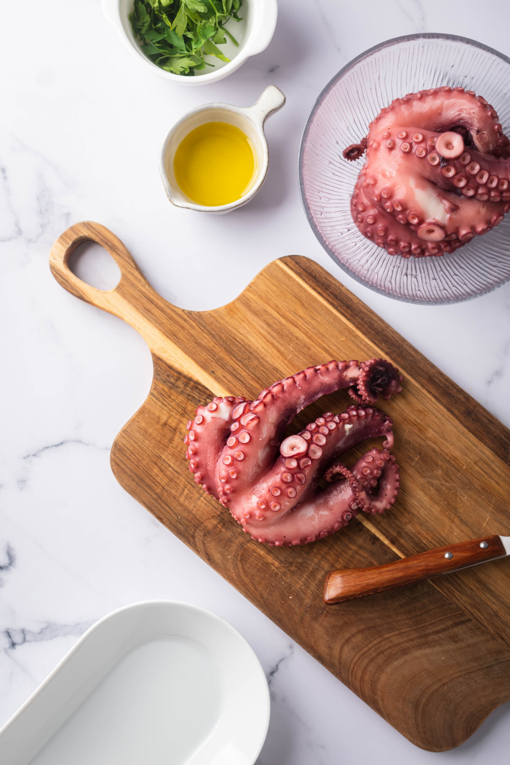 Octopus tentacles on a wooden cutting board. Behind as a glass bowl with more octopus tentacles in a bowl of melted butter next to it