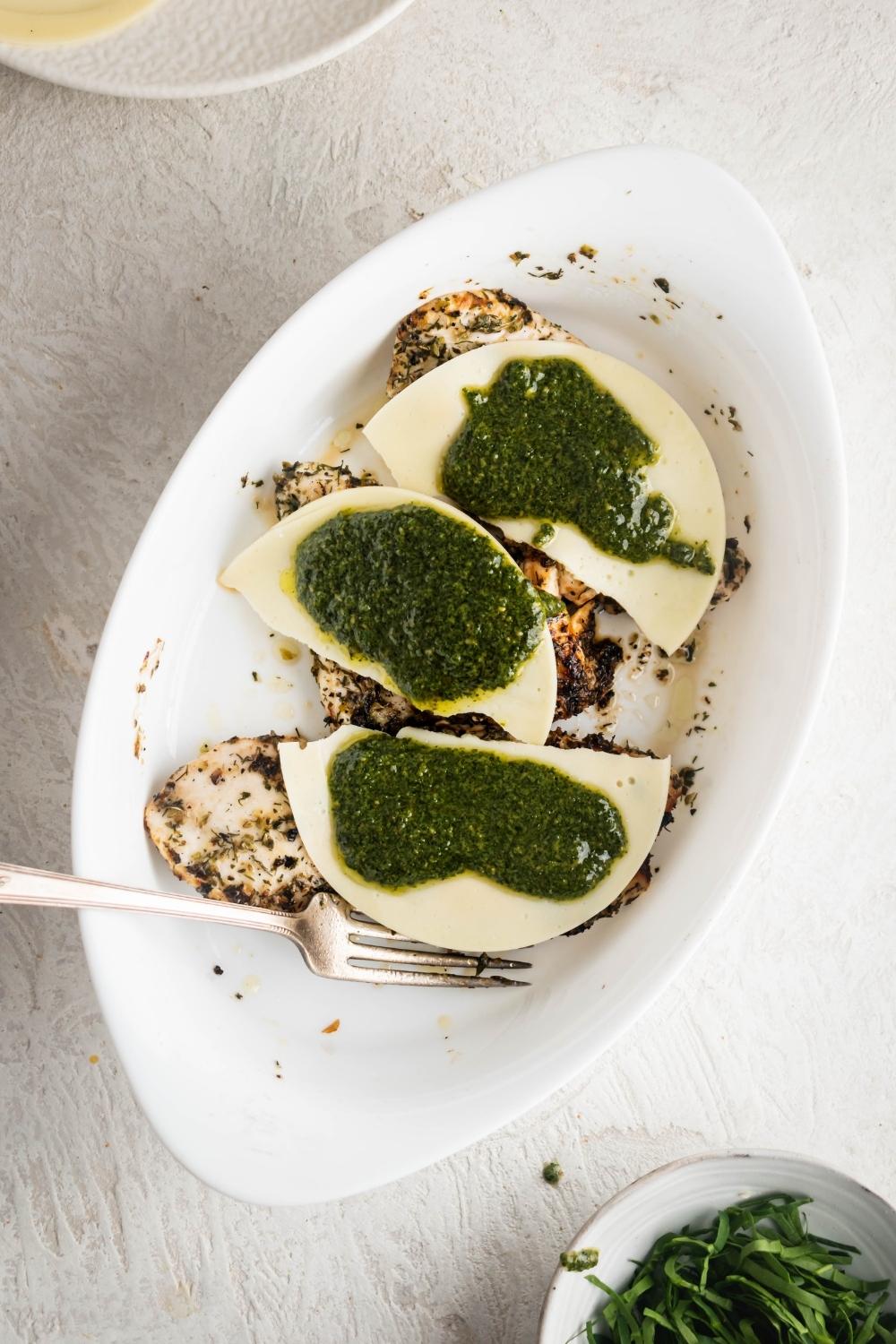 Pesto on top of three slices of mozzarella cheese on top of three chicken breasts in a white casserole dish.