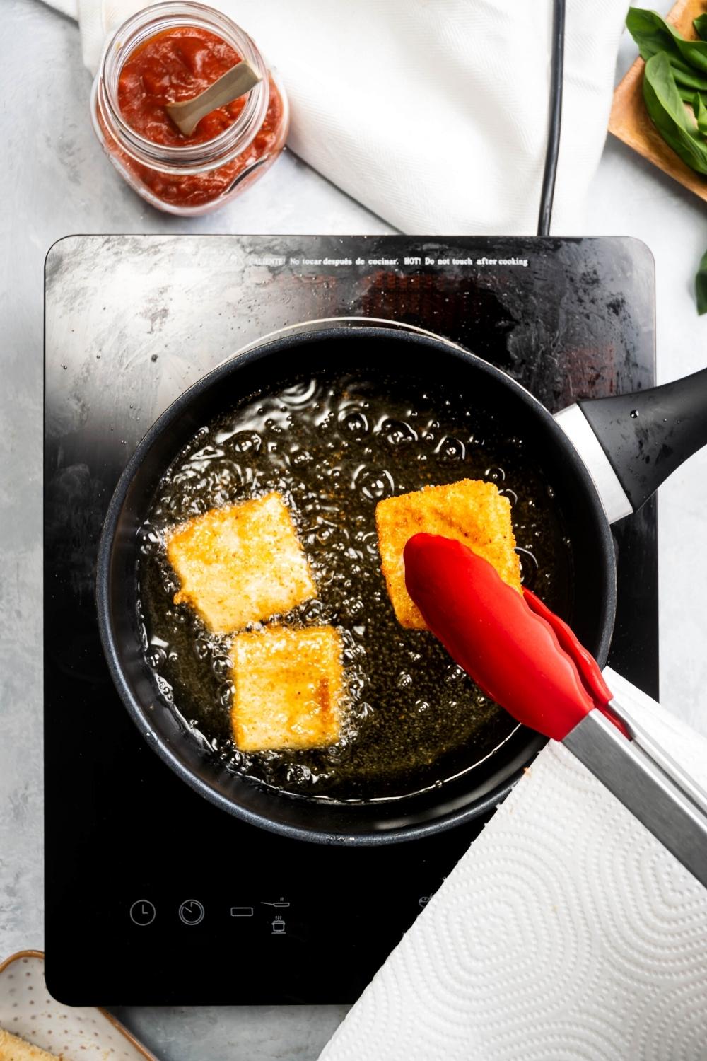 Tongs holding a piece of lasagna fritta over a pot filled with oil with two other pieces of lasagna Frida in it.