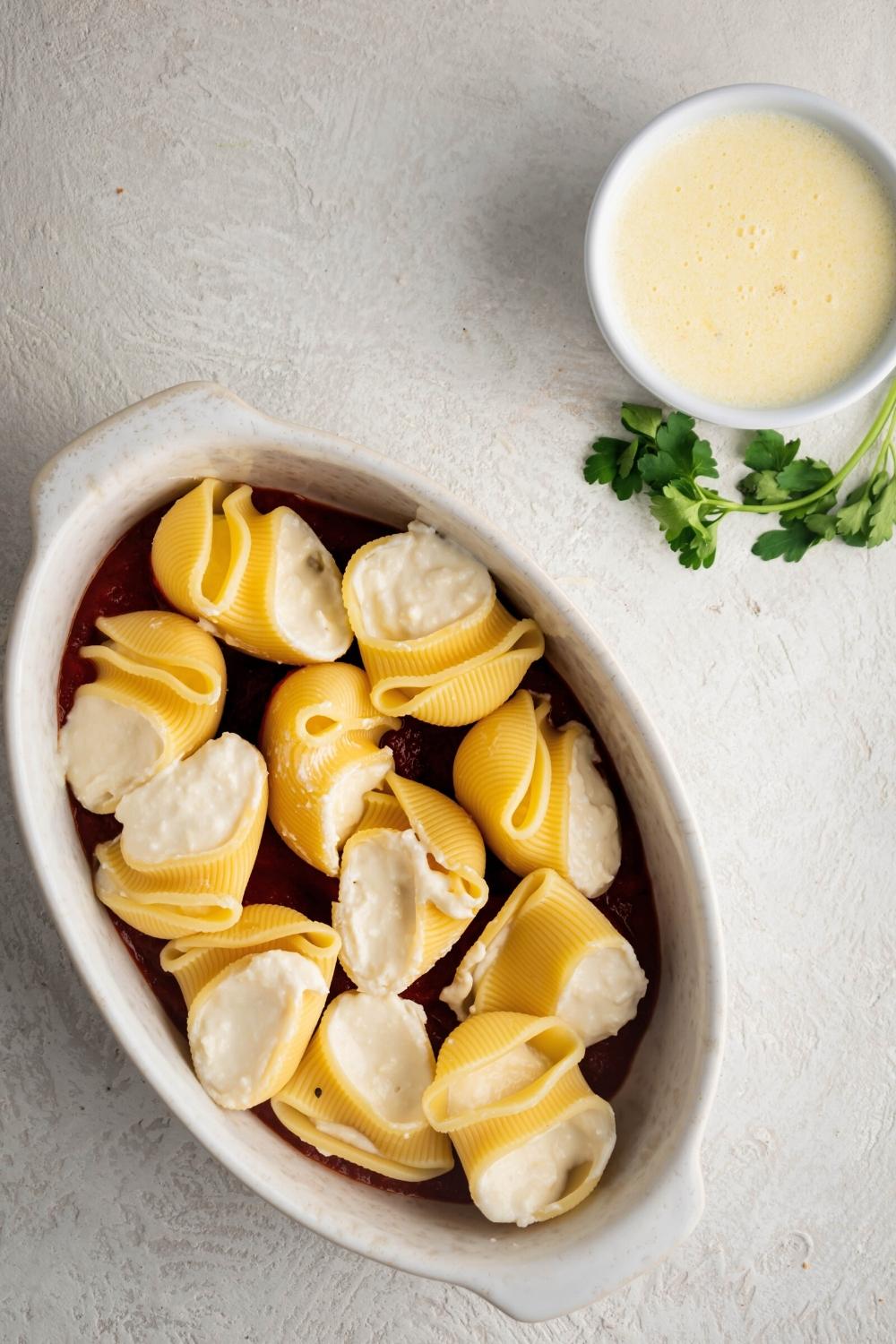 Giant cheese stuffed shells in a white baking dish. Behind it is a cup of Alfredo sauce
