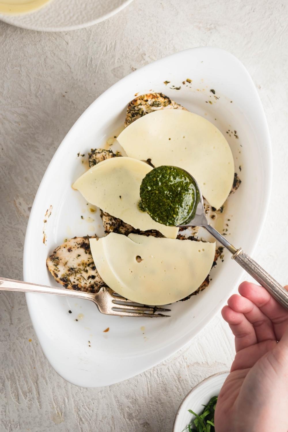 A hand holding a spoon that has pesto on it. The spoon is being held above three slices of mozzarella cheese on top of grilled chicken breasts in a white casserole dish.