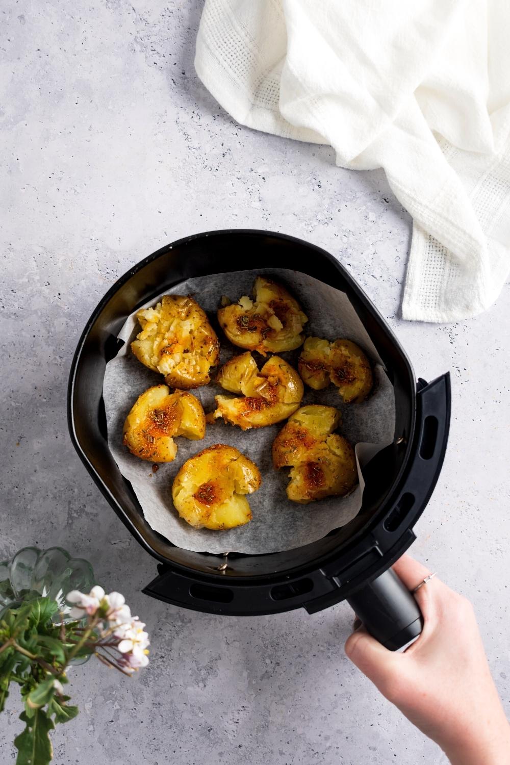 A hand holding an air fryer that has smash potatoes on top of parchment paper in the air fryer basket.