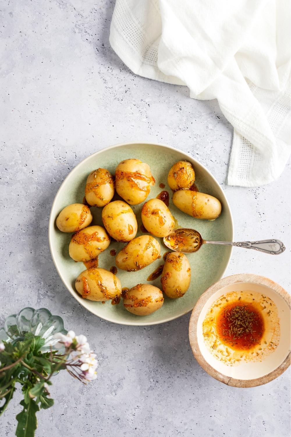 Baby potatoes with seasoning drizzled on them on top of a plate with a spoon on it. Next to the plate is a bowl with the seasoning mixture in it.