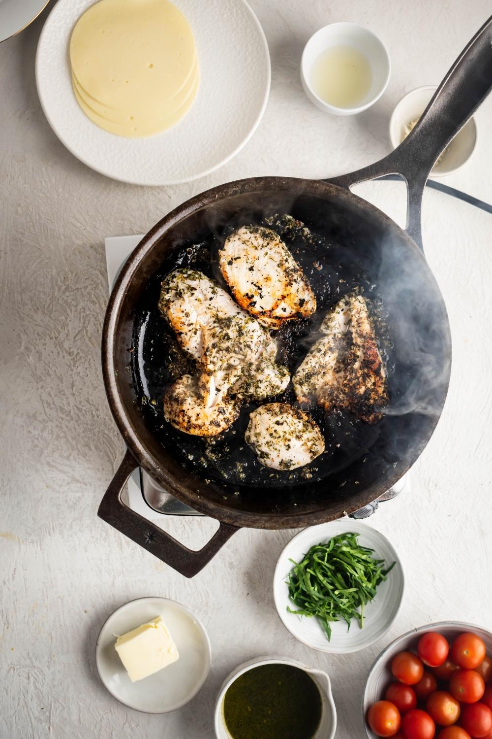 Four chicken breasts being cooked in a skillet. The chicken breasts are surrounded by a plate of mozzarella cheese, a bowl of pasta, part of a ball grape tomatoes, and a plate with a slab butter on it on a white counter.