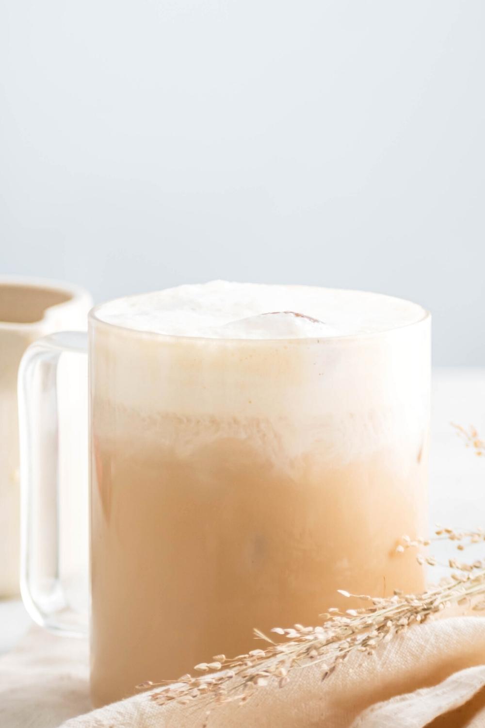 A glass filled with an iced white chocolate mocha. In front of the bottom of it is a white tablecloth and a piece of straw.