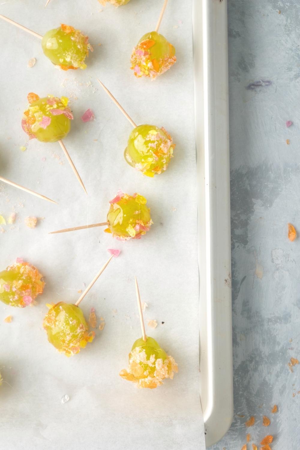 Eight candied grapes with toothpicks in them and crushed of jolly ranchers on the grapes on top piece of parchment paper on part of a baking sheet.