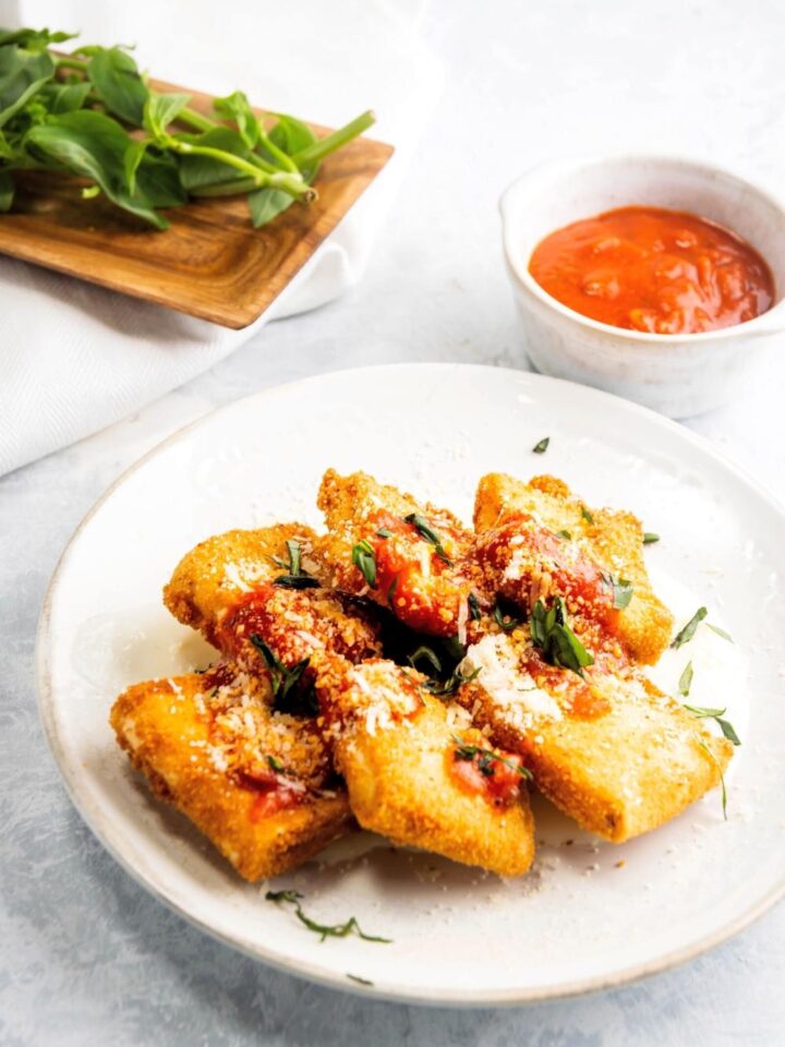 Six pieces of lasagna fritta in two lines all touching one another on a white plate. Behind it is a cup of marinara sauce.